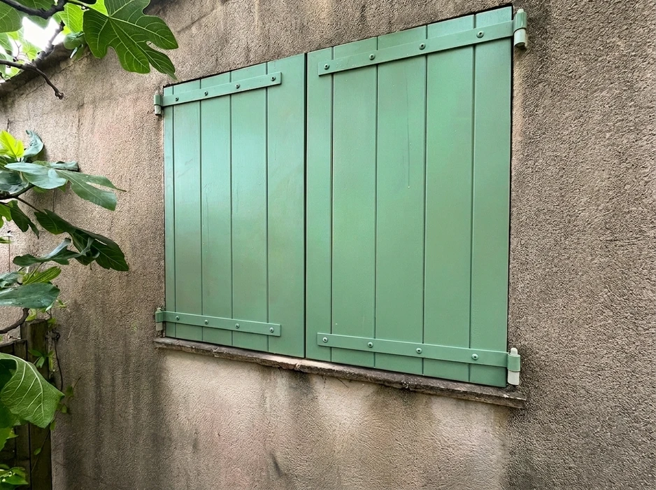 Façade extérieure de bâtiment avec fenêtre équipée d'un volet roulant blanc fermé, encadrée de deux volets battants en bois massif de teinte brune
