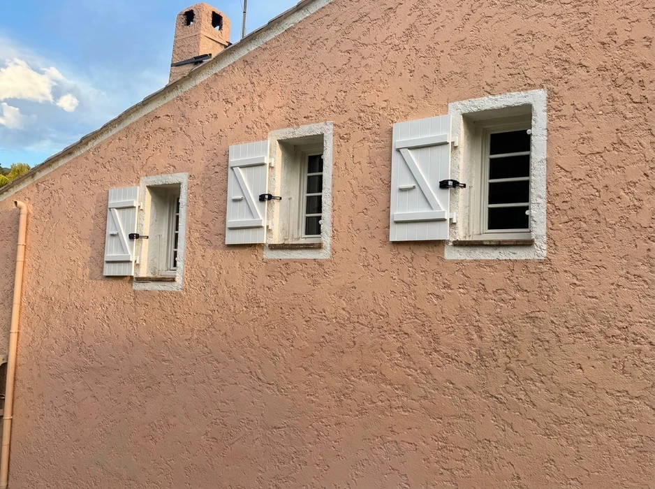 Façade extérieure de bâtiment avec fenêtre équipée d'un volet roulant blanc fermé, encadrée de deux volets battants en bois massif de teinte brune