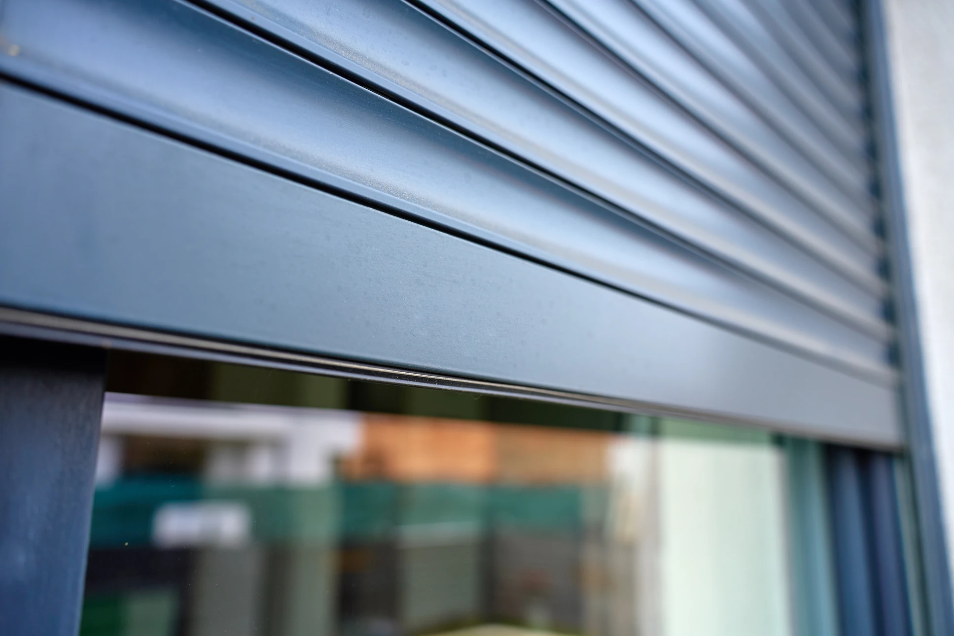 Ouvrier en combinaison rouge installant des volets roulants extérieurs sur échelle, travaux de menuiserie aluminium sur façade de bâtiment moderne sous ciel bleu